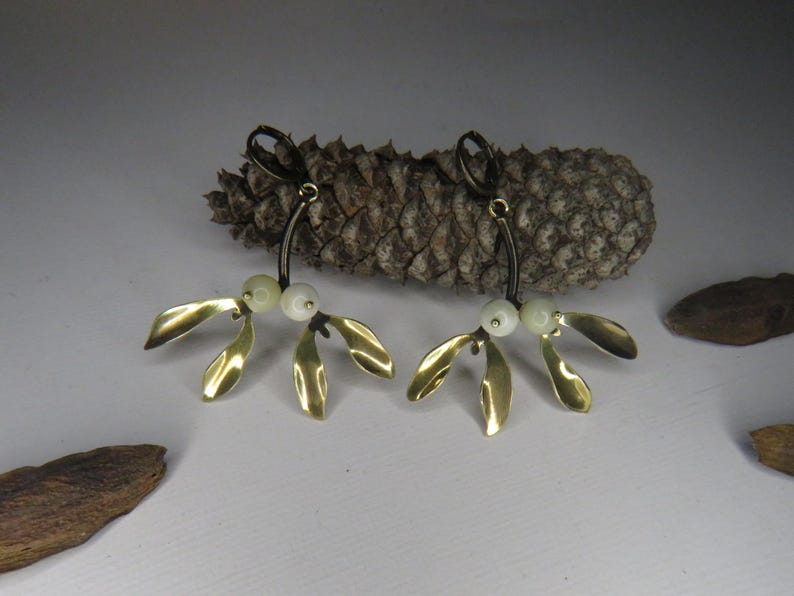 May include: A pair of gold mistletoe earrings with white pearl accents. The earrings are hanging from a brown pine cone.