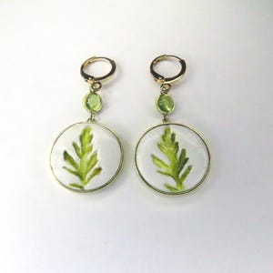 May include: A pair of gold-tone hoop earrings featuring a small round green stone accent and a larger circular white pendant. Each pendant displays a detailed green leaf illustration centered within a smooth, glossy, white circular frame.