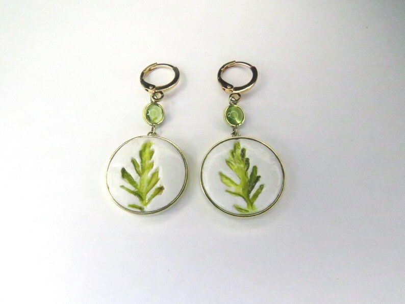 May include: A pair of gold-tone hoop earrings featuring a small round green stone accent and a larger circular white pendant. Each pendant displays a detailed green leaf illustration centered within a smooth, glossy, white circular frame.