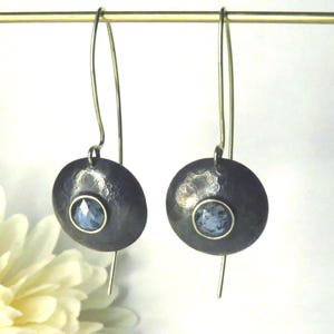 May include: A pair of dark gray, hammered metal earrings with a circular design. Each earring features a light blue gemstone set in a gold-colored bezel. The earrings hang from thin, silver-colored wires.