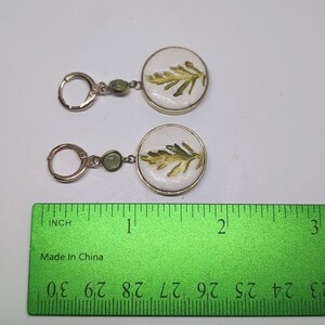 May include: A pair of gold-toned hoop earrings featuring a small green bead and a circular white pendant with a green leaf illustration. A green ruler is positioned below the earrings, showing the pendants are approximately 0.75 inches in diameter.