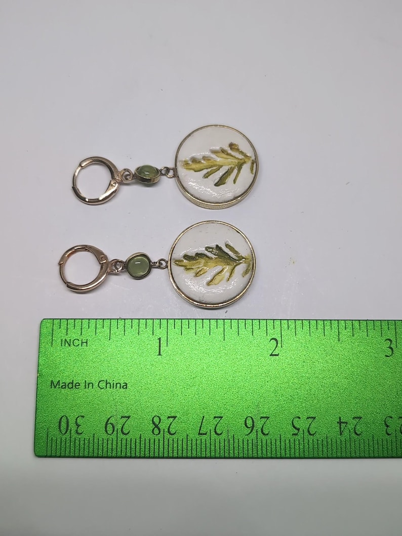 May include: A pair of gold-toned hoop earrings featuring a small green bead and a circular white pendant with a green leaf illustration. A green ruler is positioned below the earrings, showing the pendants are approximately 0.75 inches in diameter.