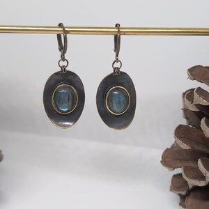 May include: A pair of dangle earrings featuring oval-shaped, dark metallic settings with a distressed finish. Each earring contains a central, smooth, iridescent blue-grey stone framed by a thin gold-toned border, suspended from a lever-back clasp.