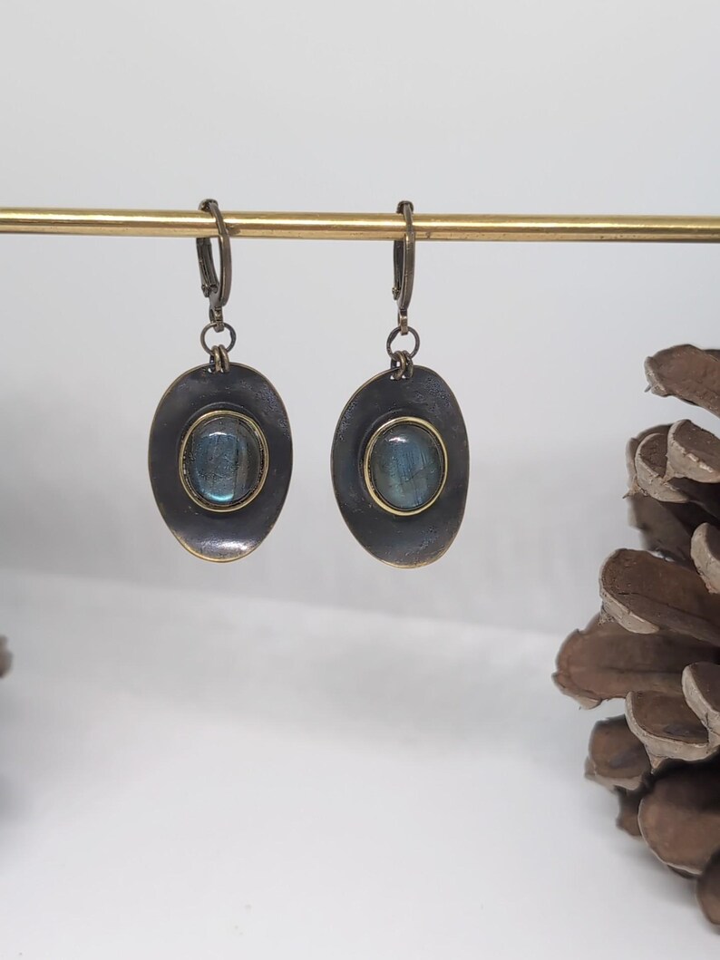 May include: A pair of dangle earrings featuring oval-shaped, dark metallic settings with a distressed finish. Each earring contains a central, smooth, iridescent blue-grey stone framed by a thin gold-toned border, suspended from a lever-back clasp.