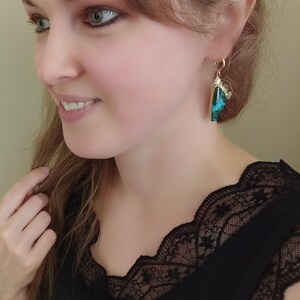 May include: A drop earring featuring a triangular-shaped piece with a vibrant teal and black marbled pattern, set in a gold-toned metal frame with a small decorative scalloped edge at the top, suspended from a simple gold-toned ear wire hook.