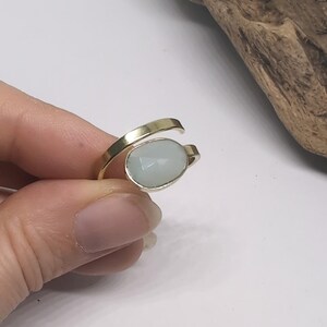 May include: A gold-toned bypass ring featuring a single oval-shaped, faceted pale blue stone set in a smooth metal bezel. The band is open at the front, with the stone positioned at one end and the plain metal band tapering at the other end.