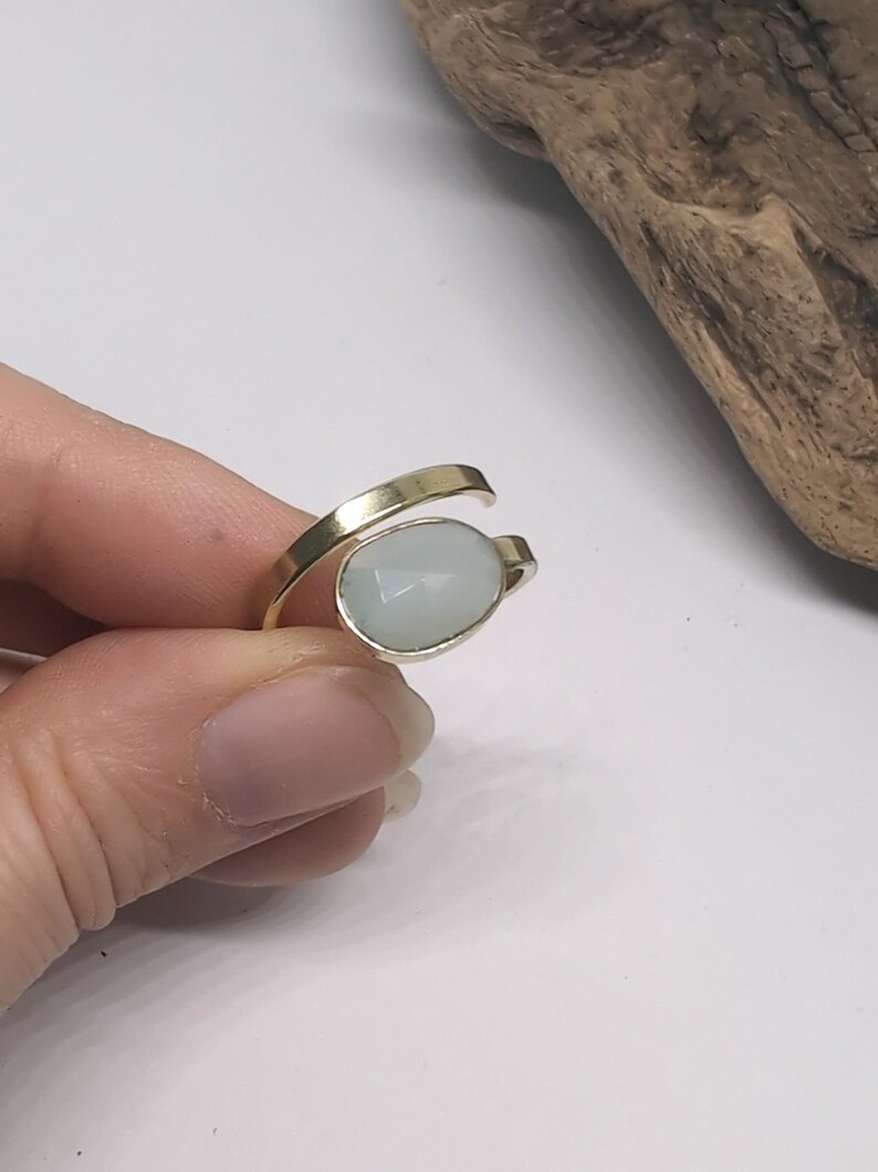 May include: A gold-toned bypass ring featuring a single oval-shaped, faceted pale blue stone set in a smooth metal bezel. The band is open at the front, with the stone positioned at one end and the plain metal band tapering at the other end.
