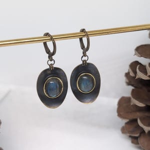 May include: A pair of drop earrings featuring oval-shaped, dark metallic settings with a distressed finish. Each earring contains a central, smooth, iridescent blue-grey stone encased in a thin gold-toned bezel, suspended from a hinged hoop clasp.