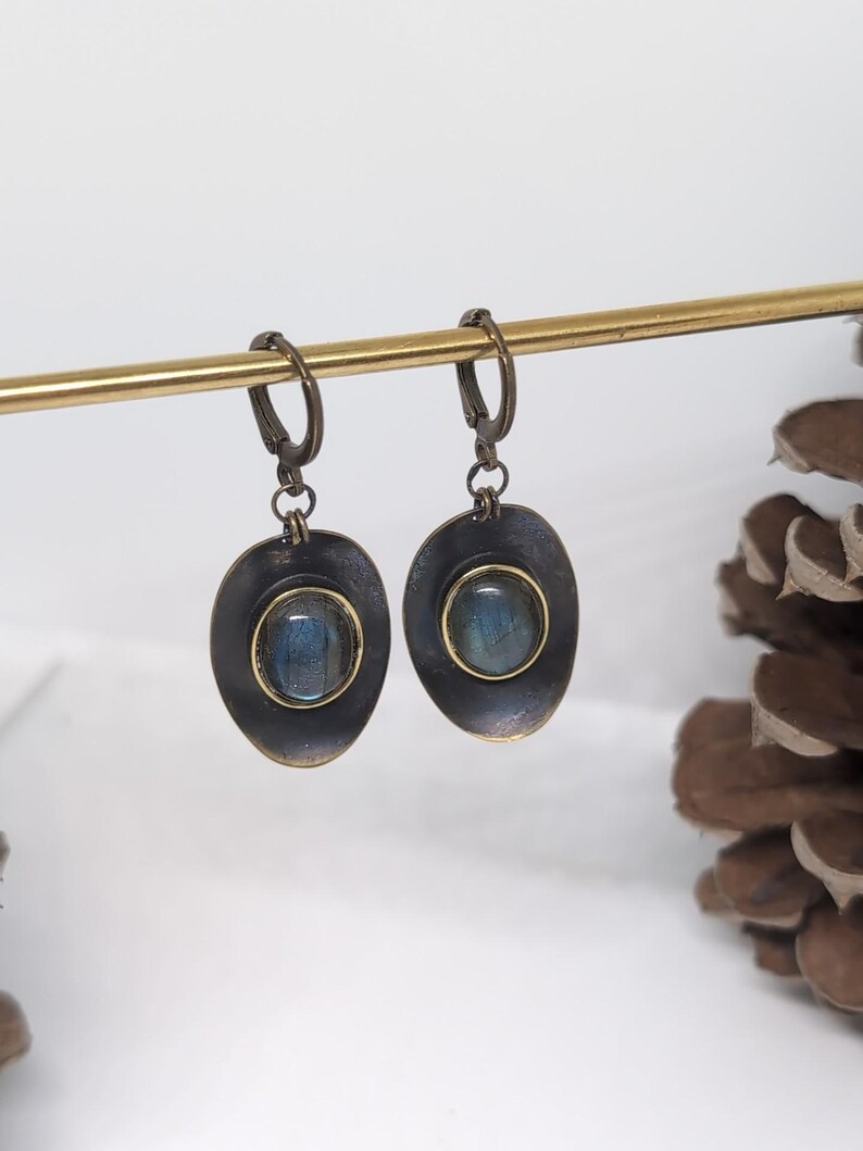 May include: A pair of drop earrings featuring oval-shaped, dark metallic settings with a distressed finish. Each earring contains a central, smooth, iridescent blue-grey stone encased in a thin gold-toned bezel, suspended from a hinged hoop clasp.