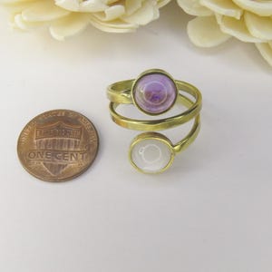 May include: A gold-toned bypass ring featuring two circular settings, one containing a light purple cabochon and the other a white cabochon, positioned next to a United States one-cent coin for scale.