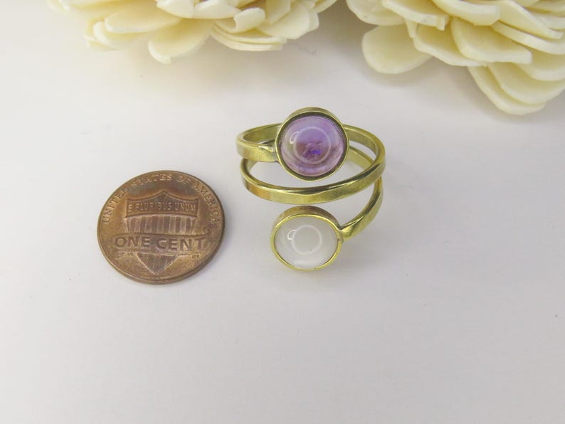 May include: A gold-toned bypass ring featuring two circular settings, one containing a light purple cabochon and the other a white cabochon, positioned next to a United States one-cent coin for scale.
