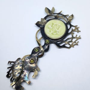 May include: A decorative metal brooch shaped like a key with an oxidized finish. It features a round, pale yellow center inlay with a relief of a frog, accented by three small green circular stones and intricate, branch-like metal cutouts.