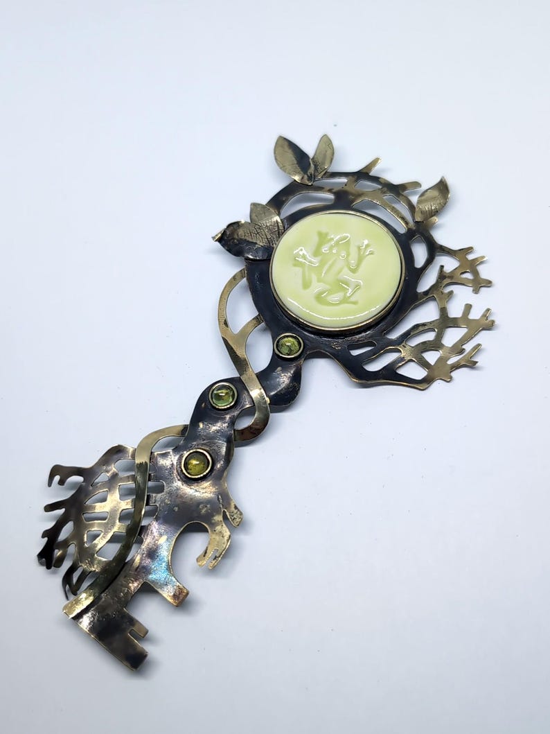 May include: A decorative metal brooch shaped like a key with an oxidized finish. It features a round, pale yellow center inlay with a relief of a frog, accented by three small green circular stones and intricate, branch-like metal cutouts.