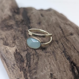 May include: A gold-toned metal ring featuring an open-ended band design with a single oval-shaped, faceted, pale blue-green stone set at one end. The ring has a smooth, polished finish and a minimalist structure that wraps partially around the finger.