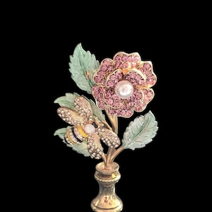 May include: A gold-toned floral brooch featuring a pink rhinestone flower with a pearl center, a rhinestone bee, and green leaves. The brooch is set against a black background, showcasing its intricate details and elegant design.