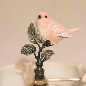 May include: A decorative lamp finial featuring a light pink bird perched on a bronze-colored branch with leaves. The bird has black eyes and is facing forward. The leaves have a glittery finish.