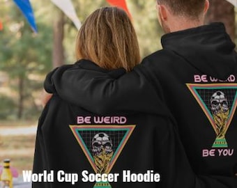 May include: Black hoodies with a soccer-themed graphic. The design features a skull, soccer ball, and trophy within a triangle, with the words "BE WEIRD" and "BE YOU". The text "World Cup Soccer Hoodie" and "Be a Goal Getter in Style!" are also visible.
