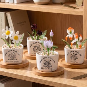 Puede incluir: Una colección de pequeñas macetas decorativas con flores de cristal, incluyendo margaritas e iris. Cada maceta cuadrada blanca se asienta sobre un posavasos de madera y presenta el texto "The Book Nook Of." con un nombre debajo.