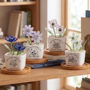 Puede incluir: Cuatro pequeñas macetas cuadradas de cerámica blanca con esculturas de flores de vidrio sobre posavasos de madera. Cada maceta está decorada con el texto "The Book Nook Of" y una ilustración con temática de libros, con diferentes nombres debajo. Las flores son azules, moradas y blancas.