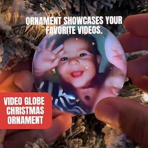 Op de afbeelding: Een heldere video globe kerstversiering met een foto van een lachend kind. De versiering wordt voor een kerstboom gehouden. Tekst op de afbeelding luidt: "Ornament showcases your favorite videos."