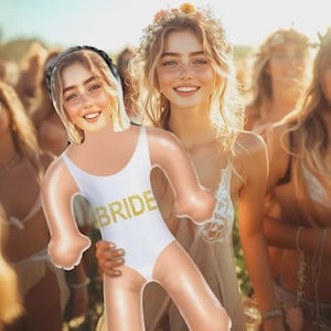 May include: A group of women smiling, one holding an inflatable doll wearing a white swimsuit with "BRIDE" in gold glitter. The doll has a black headband. The women are outdoors, some wearing flower crowns, in a sunny setting.