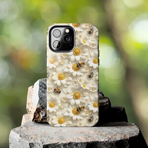 May include: White iPhone case with a floral design featuring white daisies and yellow centers.  The daisies are surrounded by small bees with black and yellow stripes.