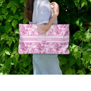 May include: A pink tote bag with a white and pink floral pattern. The bag has a white strap and the text "SARAH BRIDE" printed on the front.