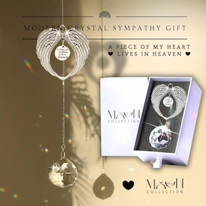 Pode incluir: Um apanhador de sol de cristal com asas de anjo prateadas com a inscrição "A piece of my heart lives in heaven". O apanhador de sol é embalado em uma caixa branca com as palavras "Moon Collection" impressas nela.