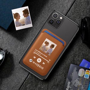 Puede incluir: Una funda de teléfono de cuero marrón con una foto de una pareja, una interfaz de reproductor de música y el texto "Line Without a Hook". La funda está adherida a un teléfono negro. También se ven una foto pequeña y tarjetas de crédito.