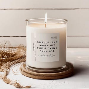 Può includere: Una candela profumata alla vaniglia accesa in un barattolo di vetro trasparente, appoggiata su un sottobicchiere di legno. L'etichetta della candela dice "SMELLS LIKE MARK HIT THE F*CKING JACKPOT" e "hand poured with Love".