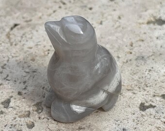 May include: A small, grey, carved bird figurine made of polished stone. The bird is sitting on a textured, light grey surface.