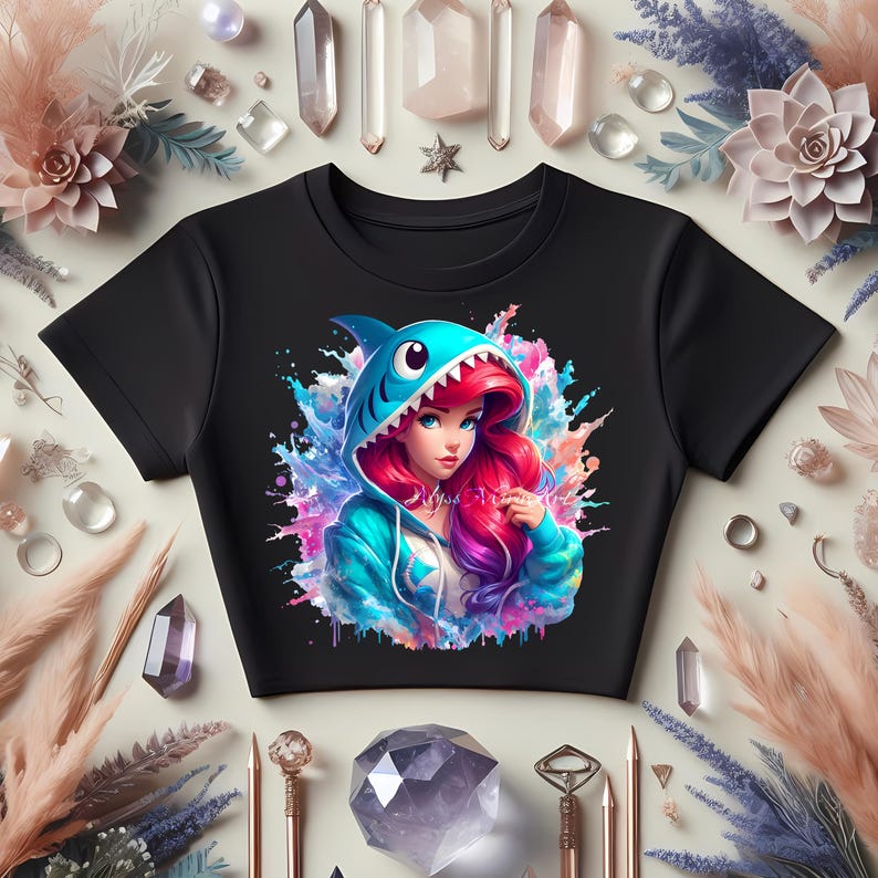 Op de afbeelding: Zwarte crop top met een kleurrijke illustratie van een vrouw met rood haar die een haaienhoodie draagt. Het ontwerp bevat spetters blauw, roze en paars. De afbeelding is omgeven door kristallen, bloemen en decoratieve elementen.