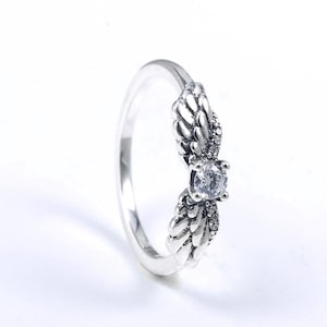 K&ouml;nnte beinhalten: Silberring mit einem Fl&uuml;geldesign auf beiden Seiten eines zentralen, klaren, runden Edelsteins. Der Ring hat eine polierte Oberfl&auml;che. Der Schmuck ist vor einem wei&szlig;en Hintergrund platziert, der die Details hervorhebt.