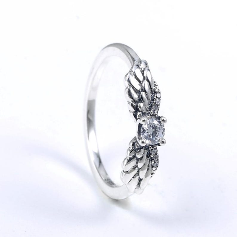 K&ouml;nnte beinhalten: Silberring mit einem Fl&uuml;geldesign auf beiden Seiten eines zentralen, klaren, runden Edelsteins. Der Ring hat eine polierte Oberfl&auml;che. Der Schmuck ist vor einem wei&szlig;en Hintergrund platziert, der die Details hervorhebt.