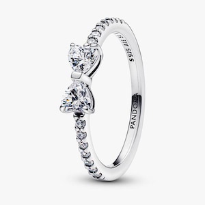 May include: A silver ring with a bow design. The ring features two clear, heart-shaped stones and a band adorned with small, clear stones. The ring is marked with "S925 ALE" and "PANDORA".