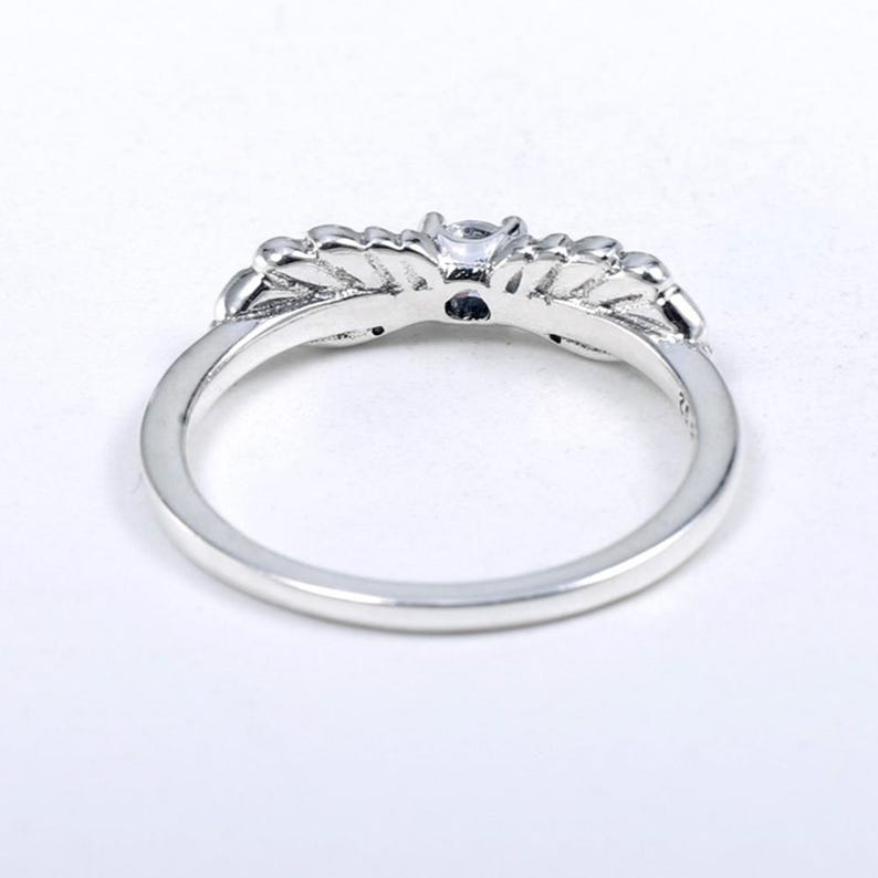 K&ouml;nnte beinhalten: Ein silberner Ring mit floralem Design. Der Ring hat einen zentralen, klaren Edelstein und kleinere Steine, die in die floralen Elemente eingesetzt sind. Das Band ist glatt und poliert und erzeugt ein zartes und elegantes Aussehen.