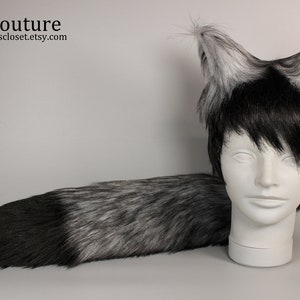 May include: A grey and black faux fur husky tail and ear set. The ears are upright with grey tips and black accents. The tail is long and fluffy, transitioning from black to grey. The set is displayed on a white mannequin head.