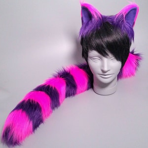 May include: A furry cat ear headband and tail set. The ears are purple with pink accents, and the tail is striped with vibrant pink and purple. The set is displayed on a white mannequin head.
