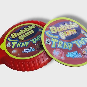 May include: Two containers of Bubble Gum Trap Tape, a tangy tropical bubble gum. The red container is oval-shaped with a ridged edge. The lid is a maroon color with yellow and blue text.