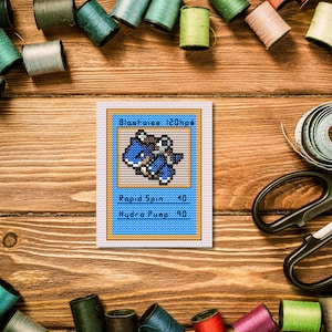 May include: A cross-stitch of a Blastoise Pokémon card, featuring a blue turtle-like creature. The card has text that reads "Blastoise 120hp", "Rapid Spin 40", and "Hydro Pump 90". The card is surrounded by spools of thread and scissors.