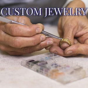 Könnte beinhalten: Ein goldener Ring, der mit einer Metallpinzette bearbeitet wird, mit dem Text CUSTOM JEWELRY oben. Eine durchsichtige Kunststoff-Sortierbox mit verschiedenen kleinen, bunten Edelsteinen befindet sich im Vordergrund für die Schmuckmontage.