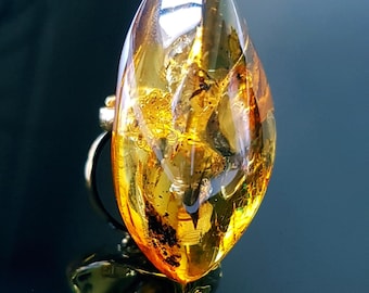 May include: A polished, oval-shaped amber gemstone ring featuring a translucent, golden-orange hue with visible organic inclusions trapped inside the resin. The smooth, rounded stone is securely mounted on a simple, thin metallic band.