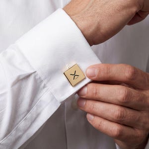 May include: Gold-colored square cufflinks with the XRP cryptocurrency symbol. The cufflinks are on a white dress shirt. The XRP symbol is black and is in the center of the square.