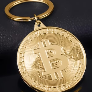May include: A gold-colored Bitcoin keychain. The Bitcoin symbol is prominently displayed on the coin-shaped pendant. The keychain has a gold ring for attaching keys. The background is a dark, textured surface.