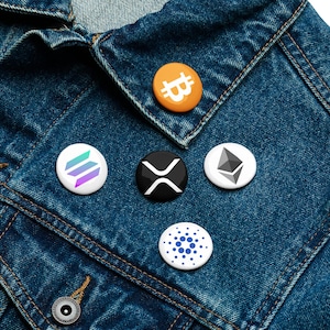 Könnte beinhalten: Eine Sammlung von fünf runden Ansteckbuttons mit Kryptowährungslogos, darunter Bitcoin, Solana, Ripple, Ethereum und Cardano. Die Buttons sind auf einer blauen Jeansjacke angeordnet und zeigen eine Vielzahl von Farben und Designs.