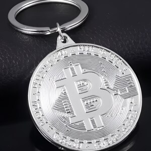 May include: Silver-toned keychain featuring a round coin-shaped charm with a raised, detailed Bitcoin symbol. The charm has a textured, circular design around the edges.