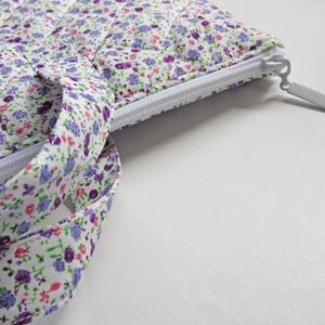 May include: A floral quilted fabric bag with a white zip and matching handles. The bag features a pattern of small purple, pink, and green flowers on a white background. The zip is open, revealing the interior.