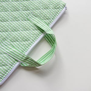 May include: A close-up of a green and white gingham patterned quilted fabric with a white zip and a green and white striped handle. The fabric is a light green colour and has a stitched, textured appearance.