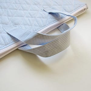 May include: A light blue quilted fabric bag with a white zip and a gingham handle. The bag has a diamond pattern and is designed for carrying items. The handle is made of a light blue and white checkered fabric.