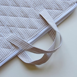 May include: Close-up of a quilted, beige and white striped fabric with a white zip and matching striped handles. The fabric has a diamond pattern created by the stitching. The handles are looped.